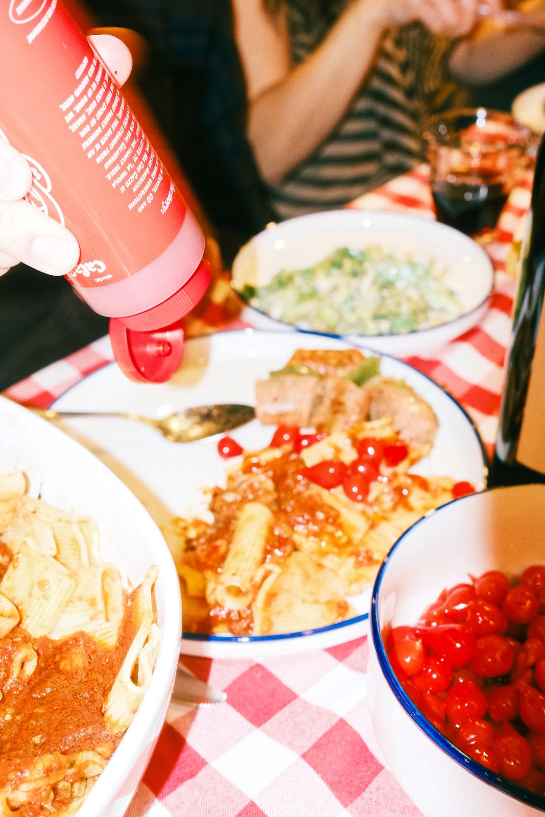 Squeezing Ma Che Cazzo over a plate of pasta, with a bowl of cherry tomatoes nearby